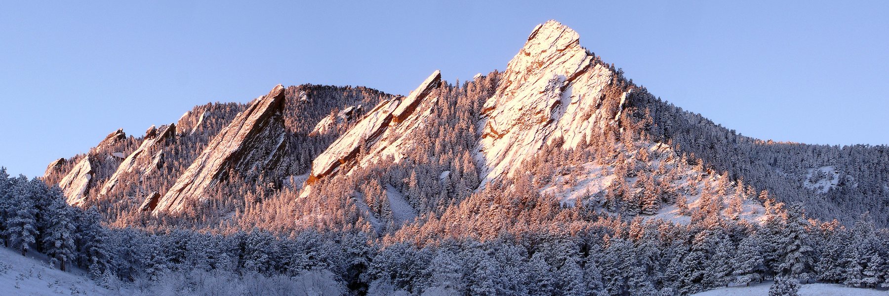 Alpenglow on the snow-dusted Flatirons above frosted pine forest under a clear blue sky